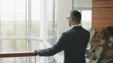 Kafkasyalı iş adamının gözlük taktığı ve gri takım elbisesinin modern ofis avlusunda panoramik manzaralı telefonla konuştuğu orta ölçekli bir fotoğraf.