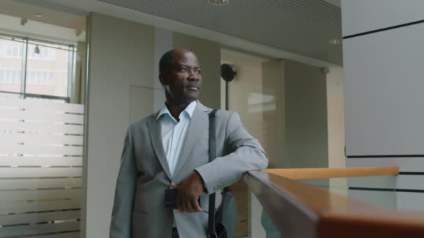 モダンなオフィスホールに立っている携帯電話とブリーフケースで灰色のスーツでアフリカ系アメリカ人の男性幹部の中型ショット — ストック動画