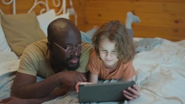 Etnik çeşitliliğe sahip baba ve oğulun tabletten film izleyerek kaliteli zaman geçirdiği orta boy bir fotoğraf.
