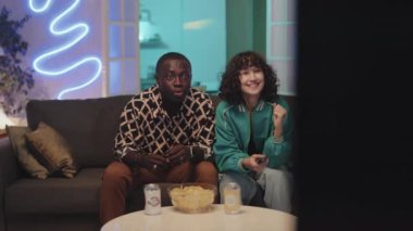 Koltukta oturup spor izleyen ve neon ışıklı odada zaferi kutlayan çok ırklı bir çift.