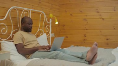 Orta boy bir Afro-Amerikan resmi. Günlük giysiler giyip yatağında dizüstü bilgisayarıyla evden uzaktan çalışıyor.