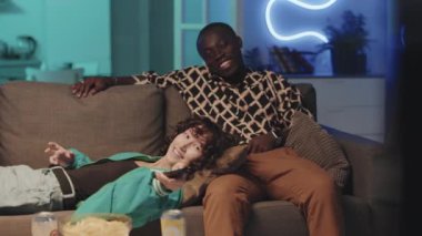 Zombi erkek oturuyor ve çift ırklı kadın uzaktan kumandayla kanalı seçiyor ve neon ışıklı odada televizyon seyrediyor.