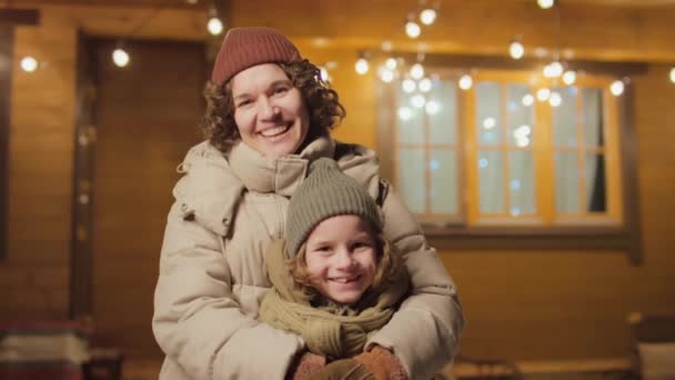 照片上快乐的妈妈和儿子穿着布拉加站在室外看着相机 背景上闪烁着圣诞彩灯 — 图库视频影像