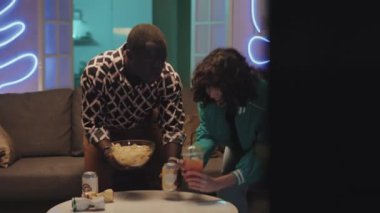 İki farklı arkadaşın aperatif ve gazoz getirip neon ışıklarıyla kanepede oturan joypad 'lerle video oyunu oynadığı tam kare çekimler.