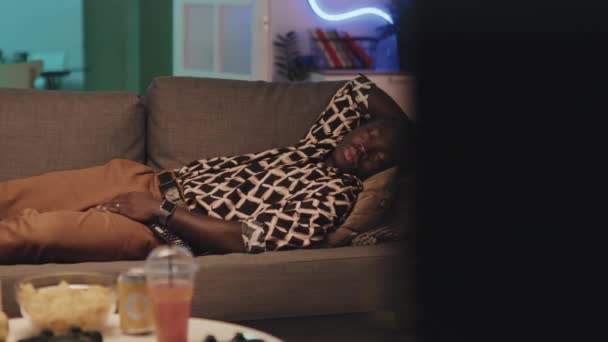 ネオンライトルームのテレビの前にソファーで昼寝をしている若いアフリカ系アメリカ人のメディアパンニングショット — ストック動画