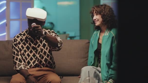 一个戴着虚拟现实耳机的非裔美国人正在学习如何与控制器玩耍 而Biracial女友则教他如何坐在有霓虹灯的沙发上 — 图库视频影像