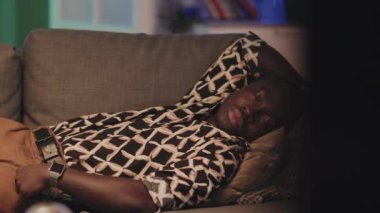 Orta boy, kareli tişörtlü ve kahverengi pantolonlu Afro-Amerikalı bir adamın oturma odasındaki kanepede uyuduğu bir fotoğraf.