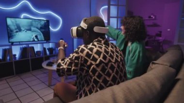 VR kulaklıklı siyahi adamın video oyunu oynarken görüntüsü. Yanında kontrolörler ve kız arkadaşları var. TV 'de izliyorlar ve neon ışıklı odada eğleniyorlar.