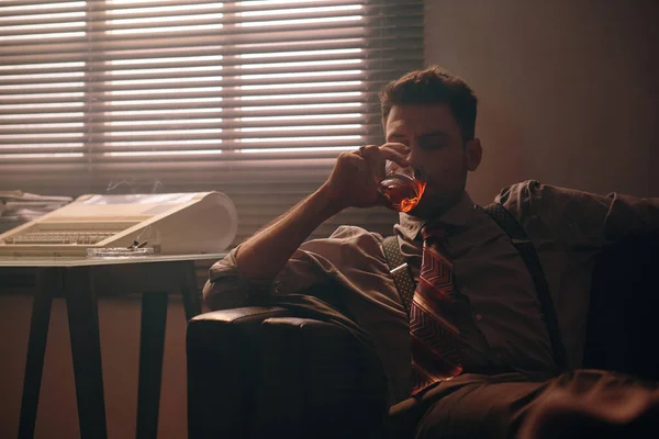 身着正式服装的年轻而安详的男子 坐在黑色皮革沙发上 靠着窗户 深色办公室的百叶窗 喝着威士忌 — 图库照片