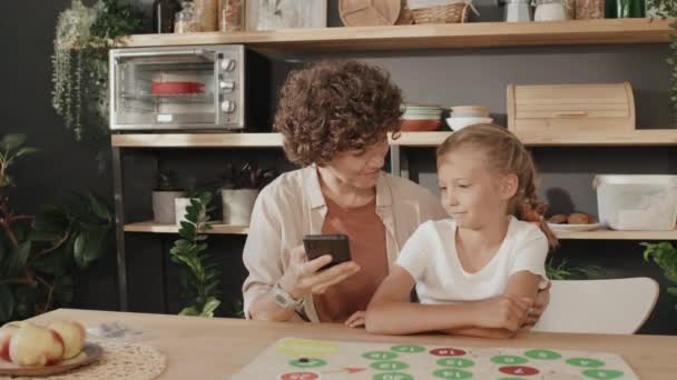 スマートフォンカメラでセルフィーを取るキッチンでテーブルに座っている現代の若い母親と彼女のプリーツの娘のスローモーション中型の肖像画 — ストック動画