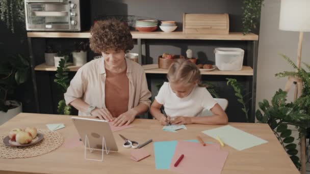 在舒适的厨房里 现代妈妈和女儿坐在餐桌旁 用蓝色和粉色的纸做折纸 动作十分缓慢 — 图库视频影像