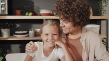 Yavaş çekim, neşeli anne ve kızının orta boy portresi. Mutfakta masada oturmuş kameraya gülümsüyor ve sarılıyorlar.