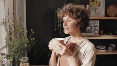 Modern beyaz kadın pencerenin önünde duruyor rahat mutfağında kahve içiyor ve manzaranın tadını çıkarıyor.