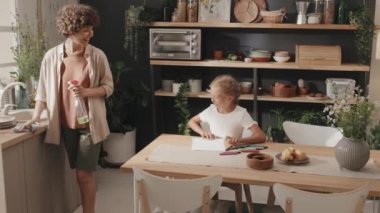 Genç kız rahat bir mutfakta masada oturuyor ve resmini annesine gösteriyor, ağır çekimde.