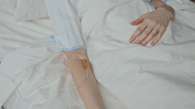 Yukarıdan bakıldığında, hastane yatağında yatan tanınmayan beyaz bir kadının kolunda sonda parçası görülüyor.