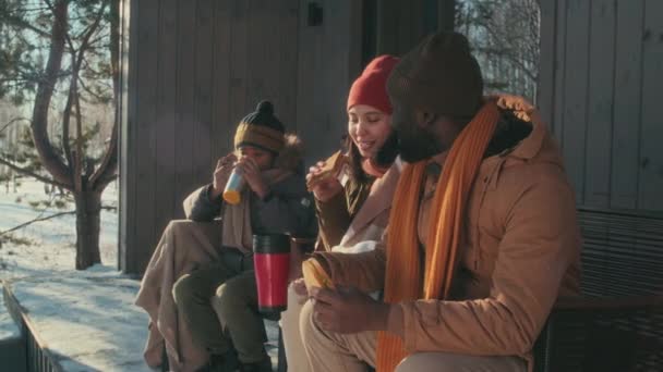 子供がポーチに座って熱いお茶を飲んで雪の冬の日にサンドイッチを食べる時間を過ごす現代人種間の家族 — ストック動画