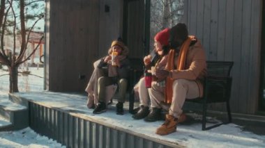 Etnik çeşitlilikte erkek, kadın ve oğulları kış günü verandada oturup sandviç yiyip sohbet ediyorlar.