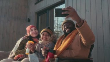 Modern etnik çeşitliliğe sahip aileler ve çocuklar kış günü verandada oturup akıllı telefondan fotoğraf çekiyorlar ve onlara bakıyorlar.