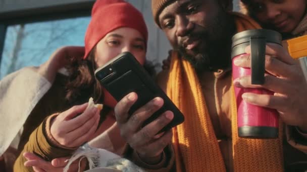 冬の日に屋外で昼食をしながら 妻と息子にインターネットでビデオを表示するスマートフォンを持っている黒人男性 — ストック動画