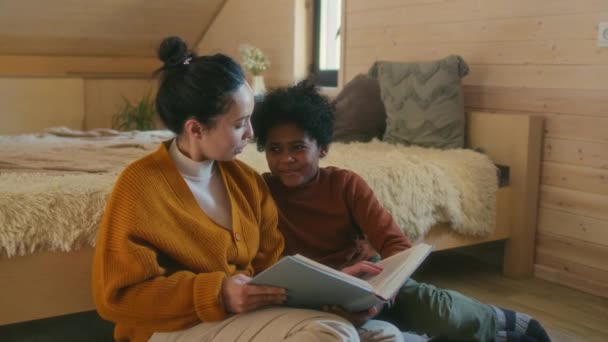 民族的に多様な母親と息子は居心地の良い寝室の読書本で床に座って冬の日を過ごしています — ストック動画