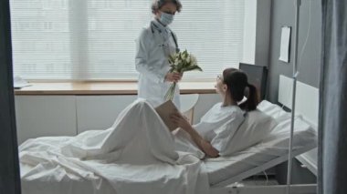 Kadın doktordan buket alan genç bir kadının hastane yatağında kitap okuması.