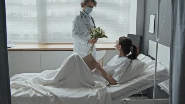 女性医師から花束を受け取る読書本で病院のベッドに横たわる若い女性のドリー — ストック動画