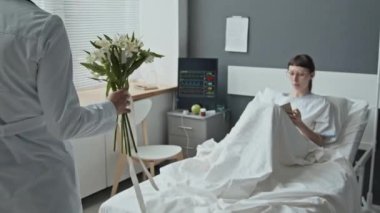 Beyaz kadın hastanın omzundan vurulup hastane yatağında yatarken tanınmayan doktorundan çiçekler alırken ve buketten notlar okurken.