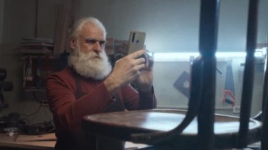 Kafkasyalı marangoz, sakallı, telefon ahşap sandalyesiyle fotoğraf çekiyor.