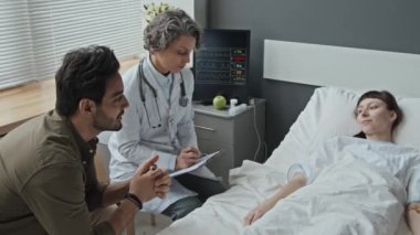 Orta boy beyaz kadın hastane yatağında uzanıp doktorla Orta Doğulu kocasının durumunu tartışıyor.