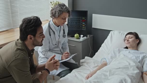 医師と中東の夫と彼女の状態を話し合って病院のベッドに横たわっている白人女性の中3ショット — ストック動画