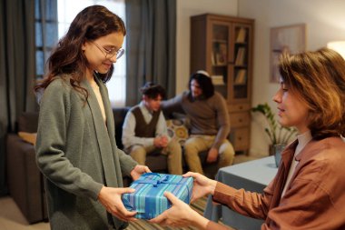 Sevimli bir kız, annesinin elinden mavi bir kağıda sarılı olarak hediye kutusunu alırken hem babasının hem de oğlunun karşısında kameranın önünde duruyor.