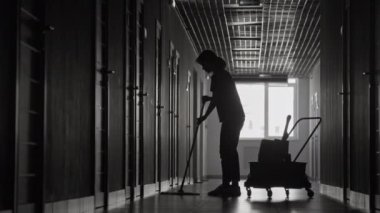 Siyah-beyaz kadın silueti. Kapıcılar üniformalı. Karanlık koridorda yerleri paspaslıyor.
