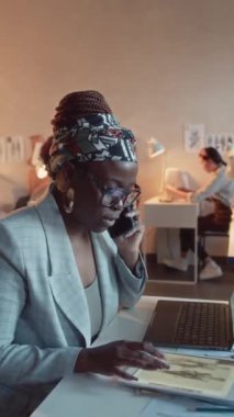 Gözlüklü ve takım elbiseli Afrikalı kadın kreatif direktörün dikey yavaşlaması telefonda konuşuyor ve atölyedeki yeni kıyafet koleksiyonunu tartışırken tabletteki çizimlere bakıyor.