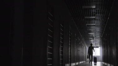 Uzun siyah beyaz erkek figürü bavul ve çanta taşıyor ve karanlık koridorda yürürken saatine bakıyor.
