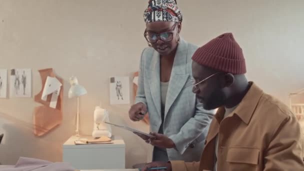 アフリカ系アメリカ人の男性ファッションデザイナーがアトリエのワークショップで新しい衣料品コレクションのスケッチに関するタブレットで黒人女性ボスに相談 — ストック動画