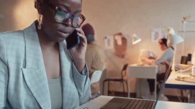 Siyah kadın iş sahibinin moda tasarımcısıyla telefonda konuşurken ve tabletteki çizimlere bakarak yeni giyim koleksiyonu tasarımını tartışırken.
