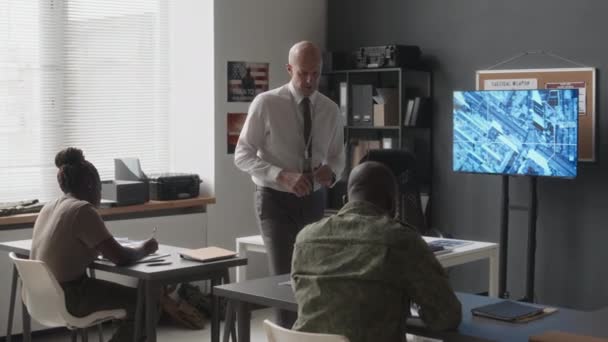 サービスアカデミーの授業中に書かれたタスクを満たす多様な認識できないキャデットを監督する教室を歩くコーカサス軍のインストラクターのフルショット — ストック動画