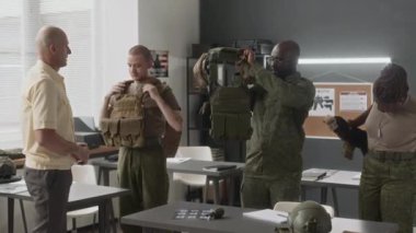 Kafkas profesörünün çeşitli öğrencilere nasıl taktik giysi giyileceğini öğrettiği orta boy bir fotoğraf. Erkek öğrenciler eğitim sırasında plaka taşıyıcıları ve taktik kemer takıyor.