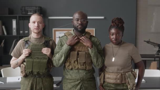 軍事アカデミーの教室に立ってカメラを見る戦術的なギアでポーズする3つの深刻な多様な女性と男性のキャデットのミディアム肖像画 — ストック動画