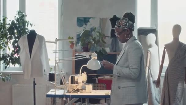 アフリカ系アメリカ人女性アトリエボスのメディアパンショット ガラスとタブレットが歩き回り 縫製機で働く多様なテールをコントロール — ストック動画