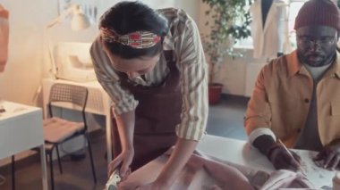 Kimliği belirlenemeyen bir kadın terzinin dikiş aletleri kullanarak tekstil keserken ve terzilik stüdyosunda elbise üretirken kumaş boşlukları hazırlarken çekimleri.