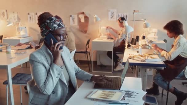 アフリカ系アメリカ人のアトリエオーナーのサイドショットが電話で話し ワークデーの間にラップトップとタブレットを使用して服のコレクションを作成するプロセスを議論 — ストック動画