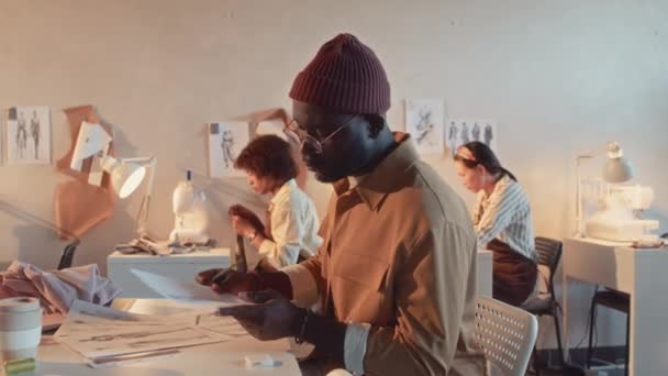 アフリカ系アメリカ人男性のスタイリッシュなファッションデザイナーのミディアムサイドショット アトリエでのワークデー中に新しいアパレルコレクションの複数のスケッチを再構築 — ストック動画