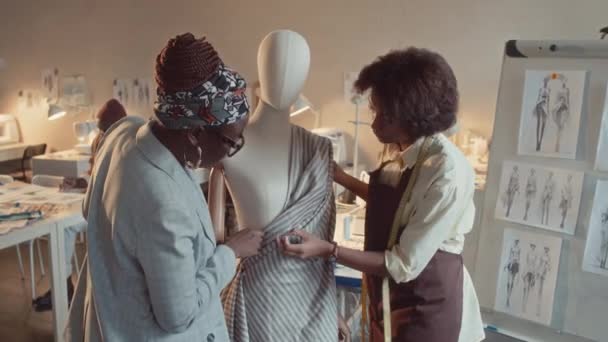 アフリカ系アメリカ人女性クリエイティブディレクターのミディアムショット ドレスを作成するプロセスを制御するタブレットと若いブラックテーラーピンクストライプファブリックをアトリエにマネキン — ストック動画