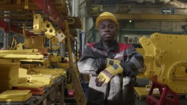 Sarı koruyucu şapkalı ve güvenlikli gülümseyen siyah işçinin orta boy portresi. Fabrikada iş günü kameraların önünde poz veriyor ve katlanır kollara bakıyor.