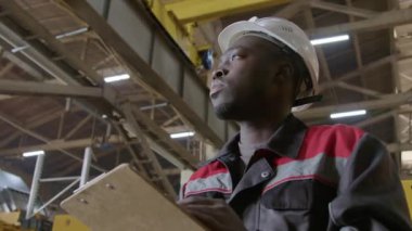Afrika kökenli Amerikalı fabrika mühendisinin düşük açılı beyaz başlık ve üniformalı makine izleme ekipmanı üretimi ve yazma verilerini fabrikada günlük raporda göster