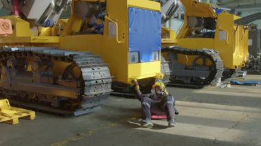 Siyah fabrika işçisinin sarı tırtıl traktör iskeletinin altından çıkıp ağır makine üretim tesisinde motor bloğunun içindeki cıvataları sıkmak için tırmanması.