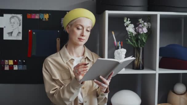 穿着黄色毛毡贝雷帽和米黄色雨衣的年轻时尚女设计师站在灯光明亮的房间里看着相机的画像 — 图库视频影像