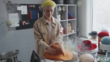 Kafkasyalı kadın zanaatkârın yeni portakalını çıkarırken orta boy bir fotoğraf. Depodan keçeli şapka çıkarıp ahşap bir mankenin üzerine koyuyor.