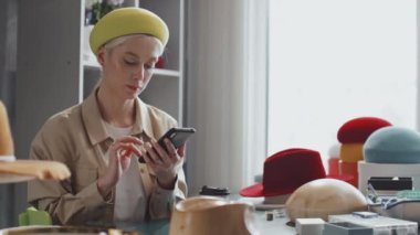 Beyaz kadın tasarımcının atölyesinde oturup telefonuna bakarken orta boy bir fotoğrafı.